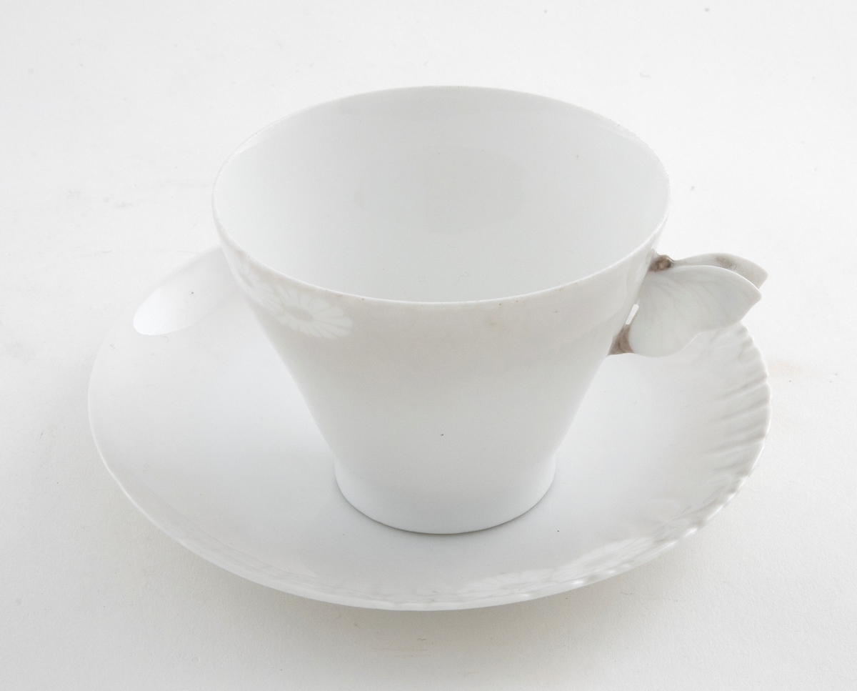 Kaffekopp med margeritter fremstilt langs kanten. Hanken er utformet som en sommerfugl. Tilhørende sirkulært fat. Langs ytterkanten er det fremstilt margeritter samt riller i lavt relieff.