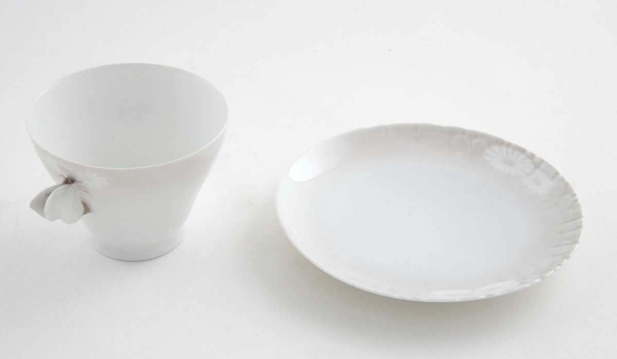 Kaffekopp med margeritter fremstilt langs kanten. Hanken er utformet som en sommerfugl. Tilhørende sirkulært fat. Langs ytterkanten er det fremstilt margeritter samt riller i lavt relieff.