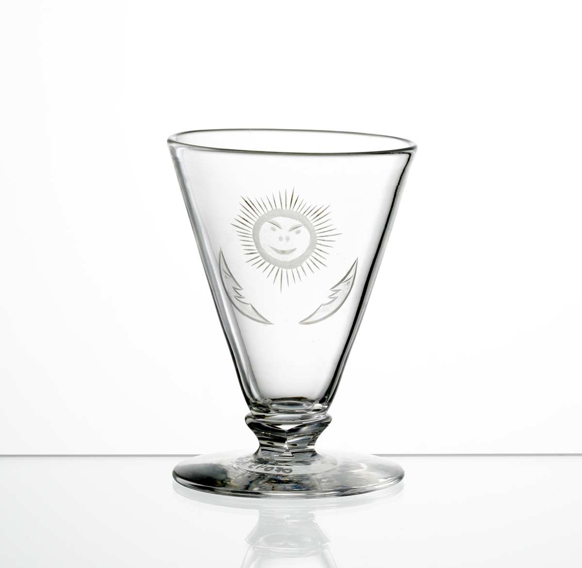 Design: Edward Hald.
Brännvinsglas med fasettslipad låg knapp mellan kupa och fot. Graverat motiv av leende sol som flankeras av två halvmånar.