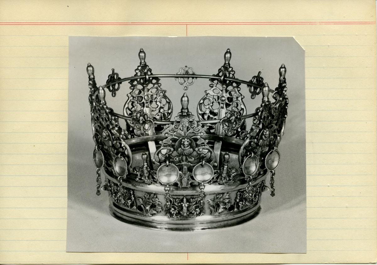 Brudkrona av förgyllt silver av Daniel Poppelman, Gävle 1770. Rikt dekorerad.
Ståndare i genombrutet arbete, 7st växtslingor med liten medaljong med renässanskvinnobild, bröstbild, i mitten. Upptill avlång topp.
På ståndarnas yttersidor hänger något nedom bildmedaljen två tunna, kupiga silverbrickor med repstav kring kanten och nertill avslutade med några länkar. Mellan ståndarnas påsatta ornament med vingad figur i genombrutet arbete; likaså vid ringens ovankant och på ringens sidor, här med rena bladornament emellan. På insidan av ringen ristat "58 1/2". Stämplar på yttrsidan av den nedre, smala ring, som håller ståndarna "G", tre kronor, "DPM" samt "M".
