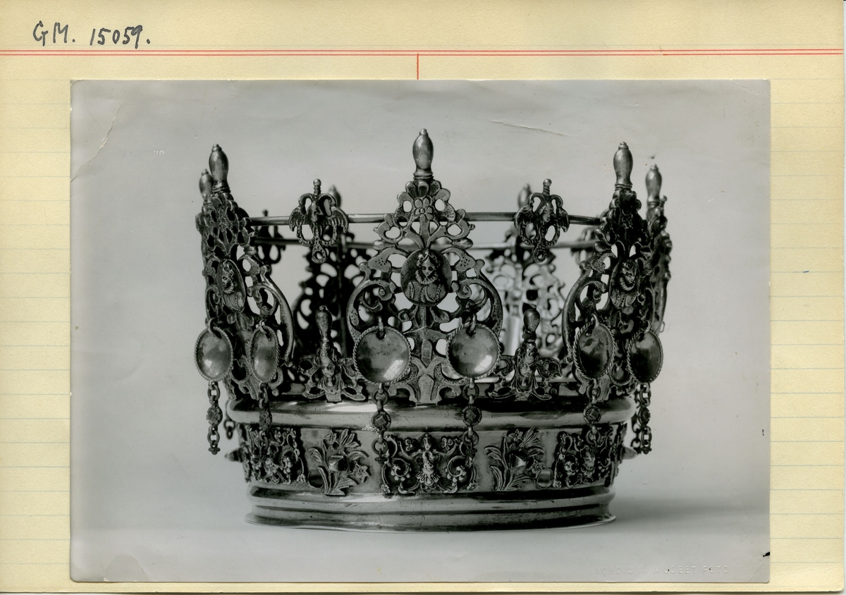 Brudkrona av förgyllt silver av Daniel Poppelman, Gävle 1770. Rikt dekorerad.
Ståndare i genombrutet arbete, 7st växtslingor med liten medaljong med renässanskvinnobild, bröstbild, i mitten. Upptill avlång topp.
På ståndarnas yttersidor hänger något nedom bildmedaljen två tunna, kupiga silverbrickor med repstav kring kanten och nertill avslutade med några länkar. Mellan ståndarnas påsatta ornament med vingad figur i genombrutet arbete; likaså vid ringens ovankant och på ringens sidor, här med rena bladornament emellan. På insidan av ringen ristat "58 1/2". Stämplar på yttrsidan av den nedre, smala ring, som håller ståndarna "G", tre kronor, "DPM" samt "M".