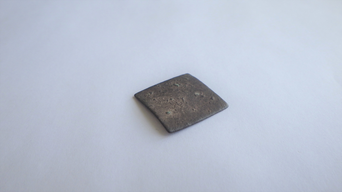 Kvadratisk metallplate med korrodert og  slitt overflate. Sider og hjørner er filt til. Det er rester av et preg på den ene flaten.