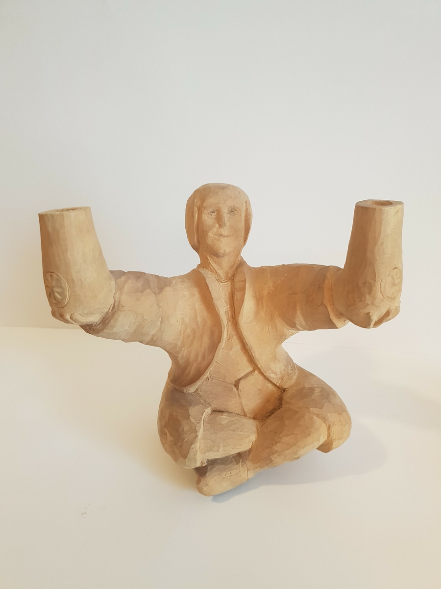 Trefigur av mann som sitter i buddah stilling og holder krukker e.l. Trolig lampet, hull på baksiden som går til hver arm\krukke. Har festeanordning på baksiden så den kan henges opp på vegg e.l. Mønster på framsiden av hver krukke, og hull i toppen.
