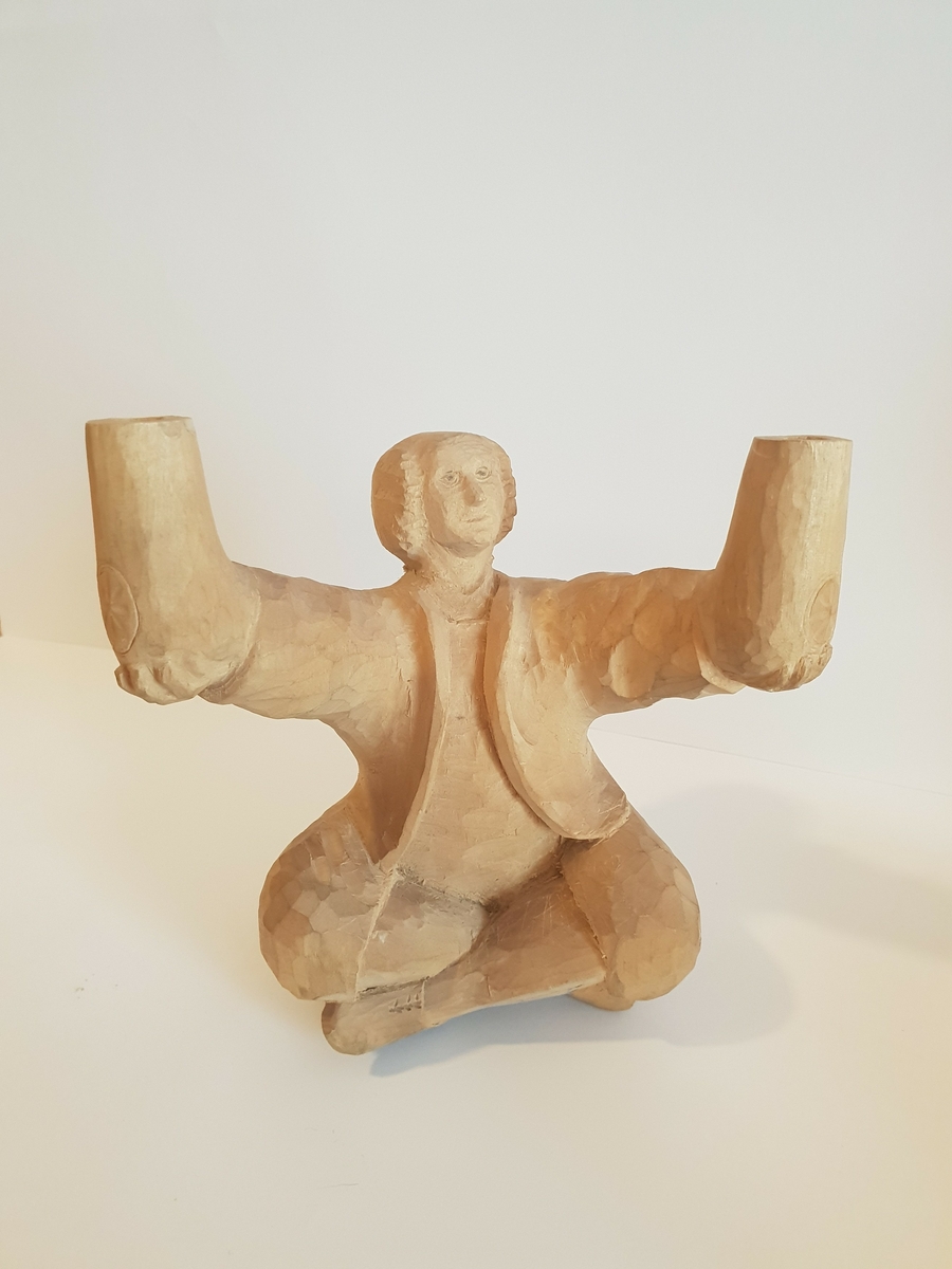 Trefigur av mann som sitter i buddah stilling og holder krukker e.l. Trolig lampet, hull på baksiden som går til hver arm\krukke. Har festeanordning på baksiden så den kan henges opp på vegg e.l. Mønster på framsiden av hver krukke, og hull i toppen.