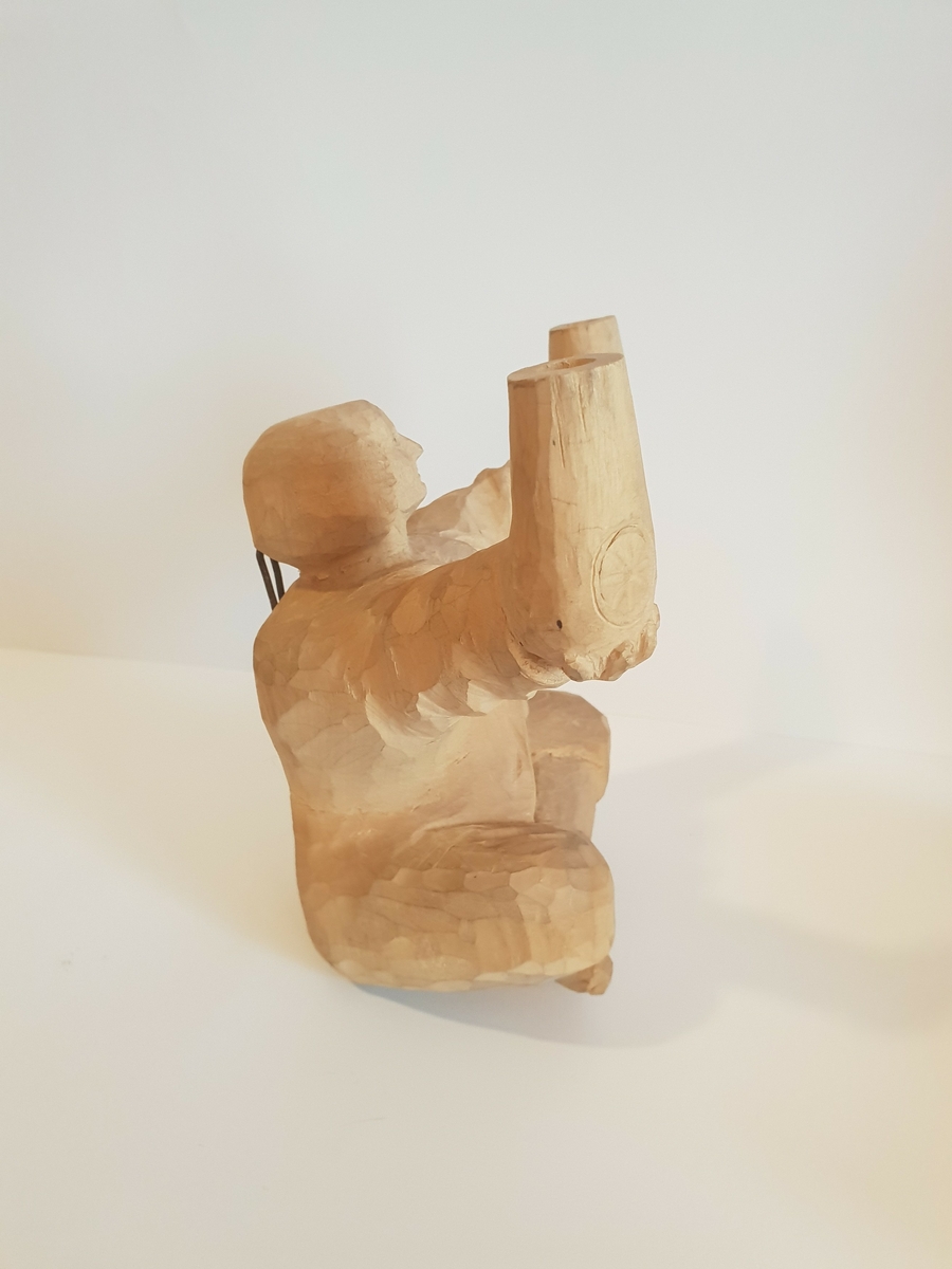 Trefigur av mann som sitter i buddah stilling og holder krukker e.l. Trolig lampet, hull på baksiden som går til hver arm\krukke. Har festeanordning på baksiden så den kan henges opp på vegg e.l. Mønster på framsiden av hver krukke, og hull i toppen.