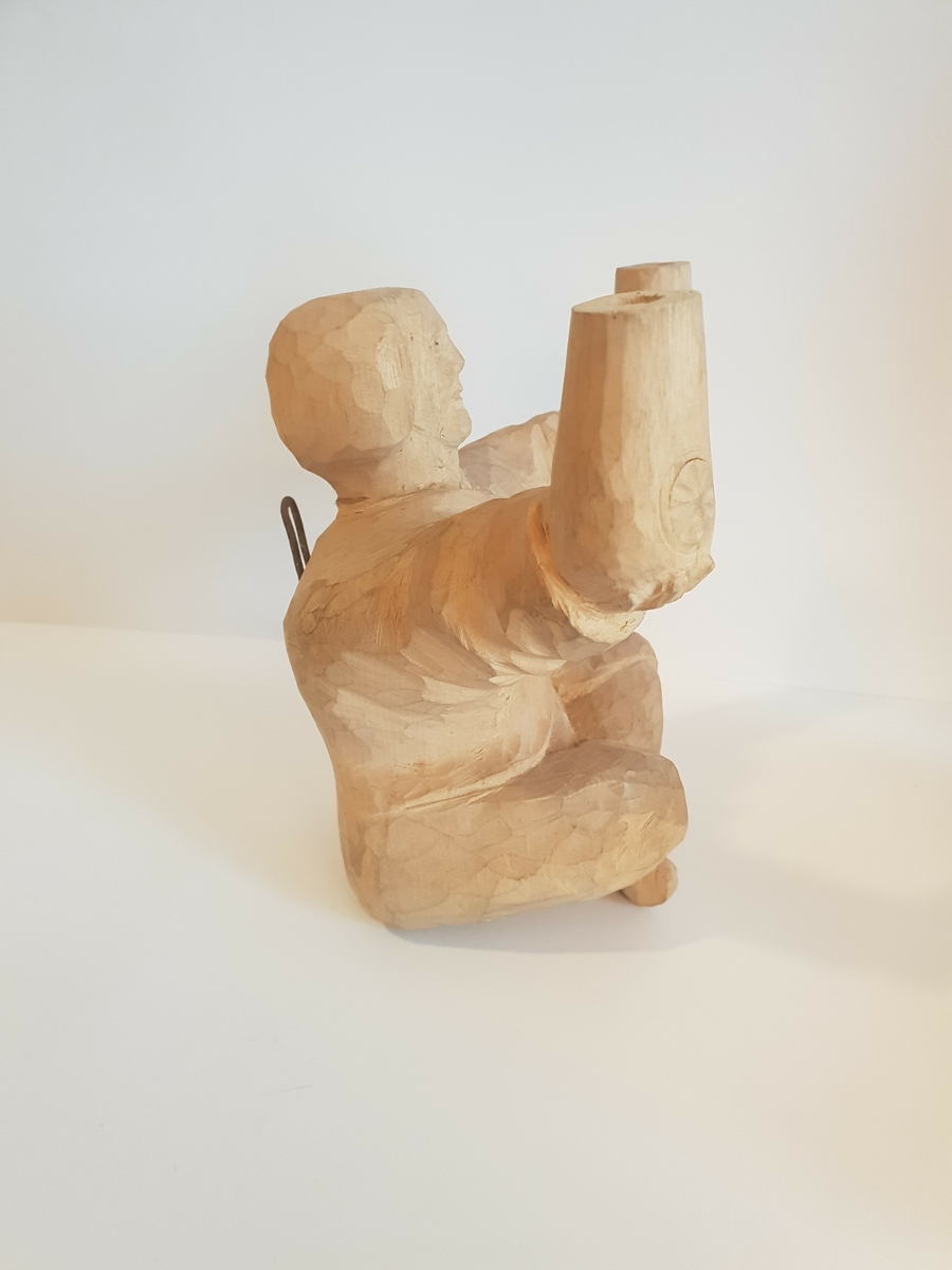Trefigur av mann som sitter i buddah stilling og holder krukker e.l. Trolig lampet, hull på baksiden som går til hver arm\krukke. Har festeanordning på baksiden så den kan henges opp på vegg e.l. Mønster på framsiden av hver krukke, og hull i toppen.