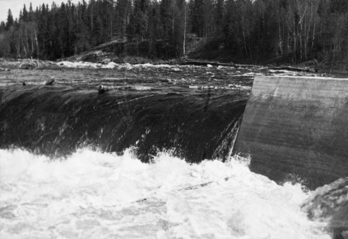 Tømmerfløting i elva Søndre Osa i Åmot kommune i Hedmark. Fotografiet er tatt våren 1936, ved den da nybygde kraftverksdammen ved Kvernfallet. I forgrunnen ser vi hvordan vannet og tømmerstokkene fløt over den avrundete damkrona. Noen av stokkene reiste seg i strømkavet under dammen, før de fløt videre. Den underliggende konstruksjonen, som vi ikke ser på dette bildet, var utført i armert betong. I 1936 var det innmeldt 383 088 tømmerstokker til fløting fra Osensjøen og tilflytende vassdrag. 28 prosent av stokkene ble levert ved Slemma, 10 prosent ved Nordre Osa, 7 prosent ved Brøa, 13 prosent ved Ena med Ulvsjøvassdraget, 7 prosent ved Tverrena og resten, 35 prosent, ble levert ved Osensjøens strender. Alt dette tømmeret. pluss noe av et kvantum på 72 506 stokker som var levert ved Søndre Osa, skulle passere den nye kraftverksdammen ved Kvernfallet. Aktørene i fløtinga var naturlig nok spente på om dammen kom til å bli et hinder der stokkene satte seg fast på eller like nedenfor dammen. Fallhøyden var heller ikke så stor at de stokkene som landet med ei av endeflatene under dammen ble påført skader. Kvernfalldammen kom ikke til å skape nevneverdige problemer for tømmerfløtinga.