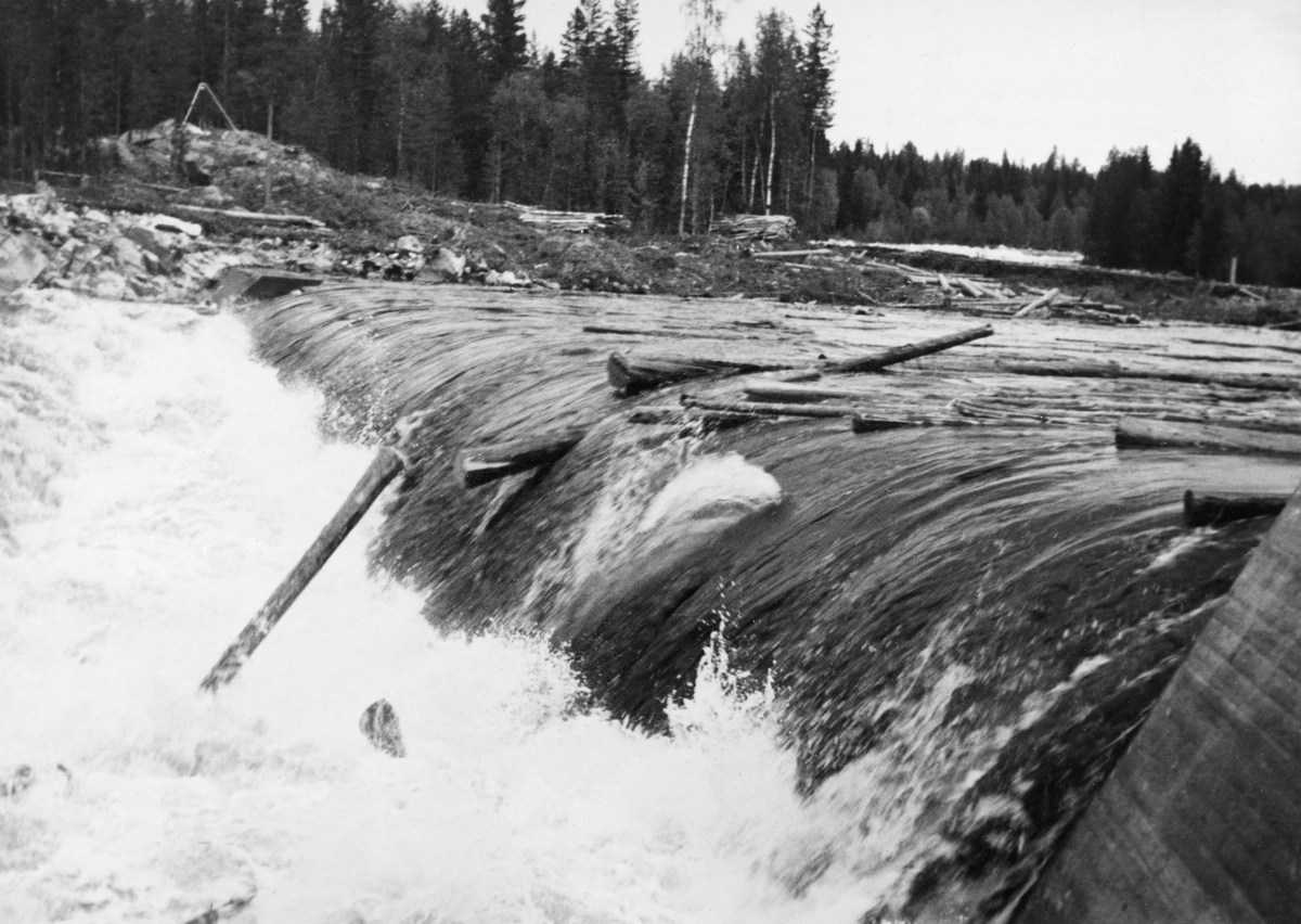 Tømmerfløting i elva Søndre Osa i Åmot kommune i Hedmark. Fotografiet er tatt våren 1936, ved den da nybygde kraftverksdammen ved Kvernfallet. I forgrunnen ser vi hvordan vannet og tømmerstokkene fløt over den avrundete damkrona. Noen av stokkene reiste seg i strømkavet under dammen, før de fløt videre. Den underliggende konstruksjonen, som vi ikke ser på dette bildet, var utført i armert betong. I 1936 var det innmeldt 383 088 tømmerstokker til fløting fra Osensjøen og tilflytende vassdrag. 28 prosent av stokkene ble levert ved Slemma, 10 prosent ved Nordre Osa, 7 prosent ved Brøa, 13 prosent ved Ena med Ulvsjøvassdraget, 7 prosent ved Tverrena og resten, 35 prosent, ble levert ved Osensjøens strender. Alt dette tømmeret. pluss noe av et kvantum på 72 506 stokker som var levert ved Søndre Osa, skulle passere den nye kraftverksdammen ved Kvernfallet. Aktørene i fløtinga var naturlig nok spente på om dammen kom til å bli et hinder der stokkene satte seg fast på eller like nedenfor dammen. Fallhøyden var heller ikke så stor at de stokkene som landet med ei av endeflatene under dammen ble påført skader. Kvernfalldammen kom ikke til å skape nevneverdige problemer for tømmerfløtinga.