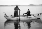 Systrarna Ester och Ingeborg Karlsson provar fiskelyckan på Sandfjärden i Sankt Anna. Bygdefotografen Emil Durling förevigade tillfället och han lät av någon anledning sin katt och hund följa med i båten. Ester var under en tid hushållerska hos Emil Durling och är även känd som Ester Bölja Bergström efter taget tillnamn och giftermål.