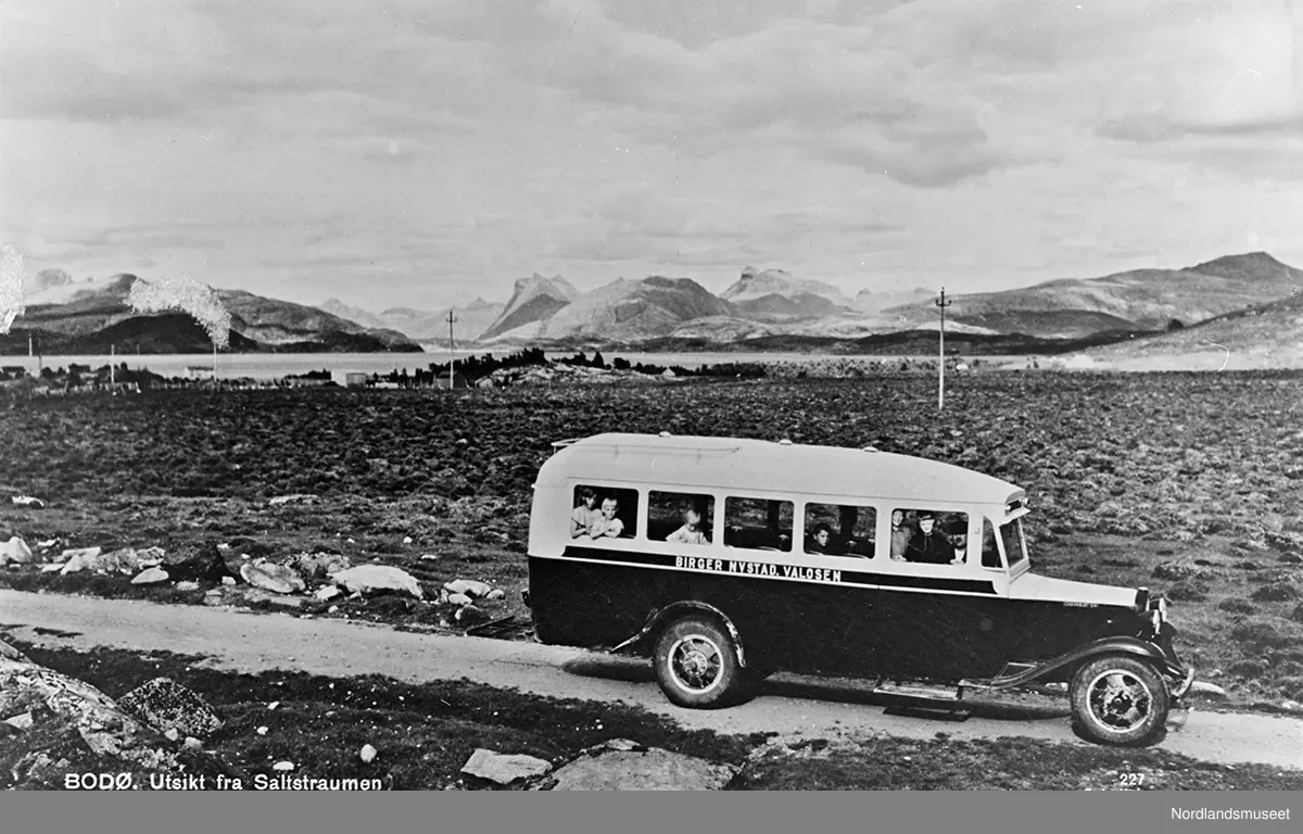 Saltstraumen, eldre buss, flere personer i bussen. På bussen står det: "Birger Nystad, Valosen". I bakgrunnen noen bolighus. I bakgrunnen Steigtind, Storviktindene og Hegmotind.