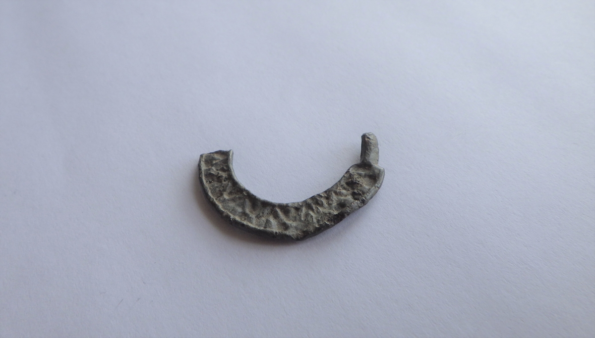 MALV: DIAM. 27  RAMMENS B. 5 MM, BÅNDFORMET. DEN ENE SIDEN HAR DEKOR, MEN VANSKELIGÅ TYDE, STØPT. SER UT SOM DEFORMERTE BOKSTAVER. LIGNER SPENNEN FRA LUND- BLOMQUIST L. 1947: BILD 22, 5. DATO: 1300-TALLET. 