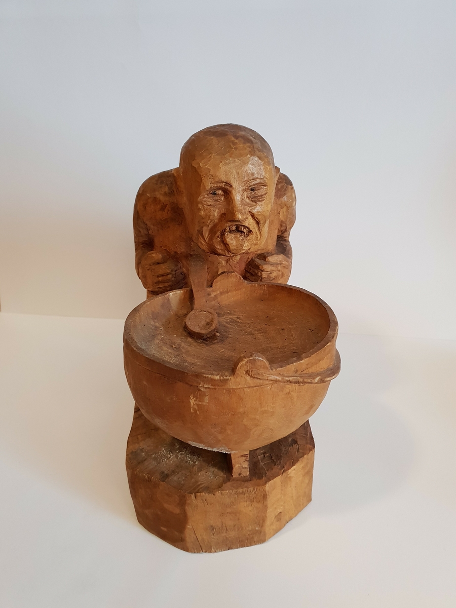 Trefigur av mann/troll? Som sitter med en gryte med en skje i. Mannen/troller henger ut med tunga.