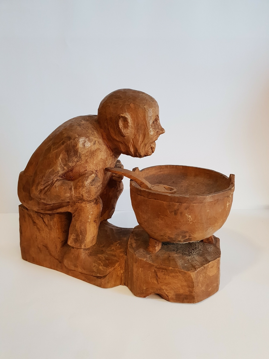 Trefigur av mann/troll? Som sitter med en gryte med en skje i. Mannen/troller henger ut med tunga.