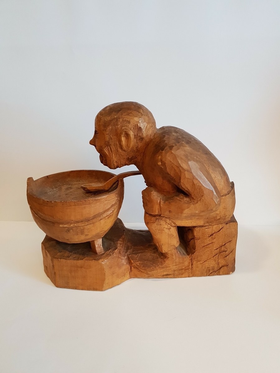 Trefigur av mann/troll? Som sitter med en gryte med en skje i. Mannen/troller henger ut med tunga.