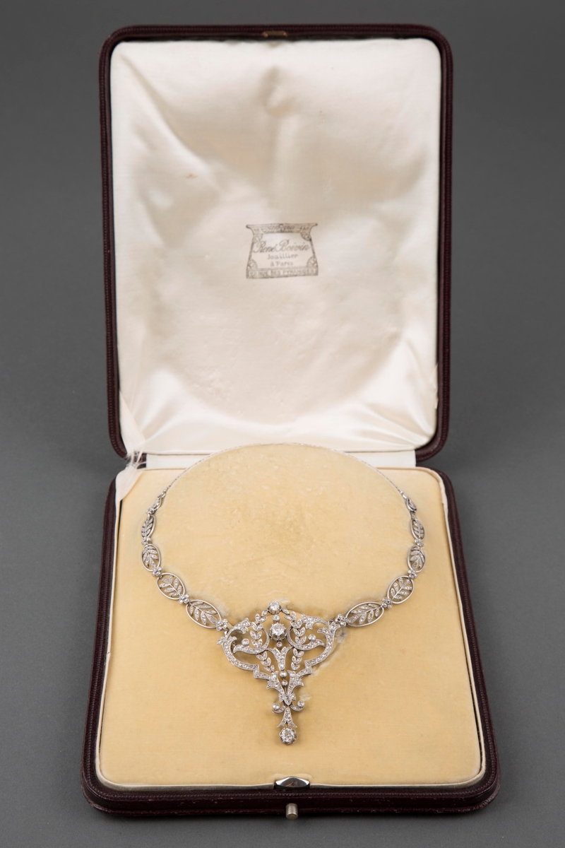 Halssmykke i platina og diamanter i originaleske. Kjedet består av ovaler med bladranker fylt med diamanter og små diamantblomster imellom. Anhenget er i hjerteform med blomster- og bladornamenter fylt med diamanter, samt runde innfattede diamanter i forskjellig størrelse.