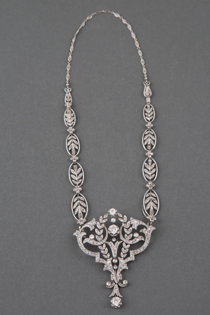 Halssmykke i platina og diamanter i originaleske. Kjedet består av ovaler med bladranker fylt med diamanter og små diamantblomster imellom. Anhenget er i hjerteform med blomster- og bladornamenter fylt med diamanter, samt runde innfattede diamanter i forskjellig størrelse.