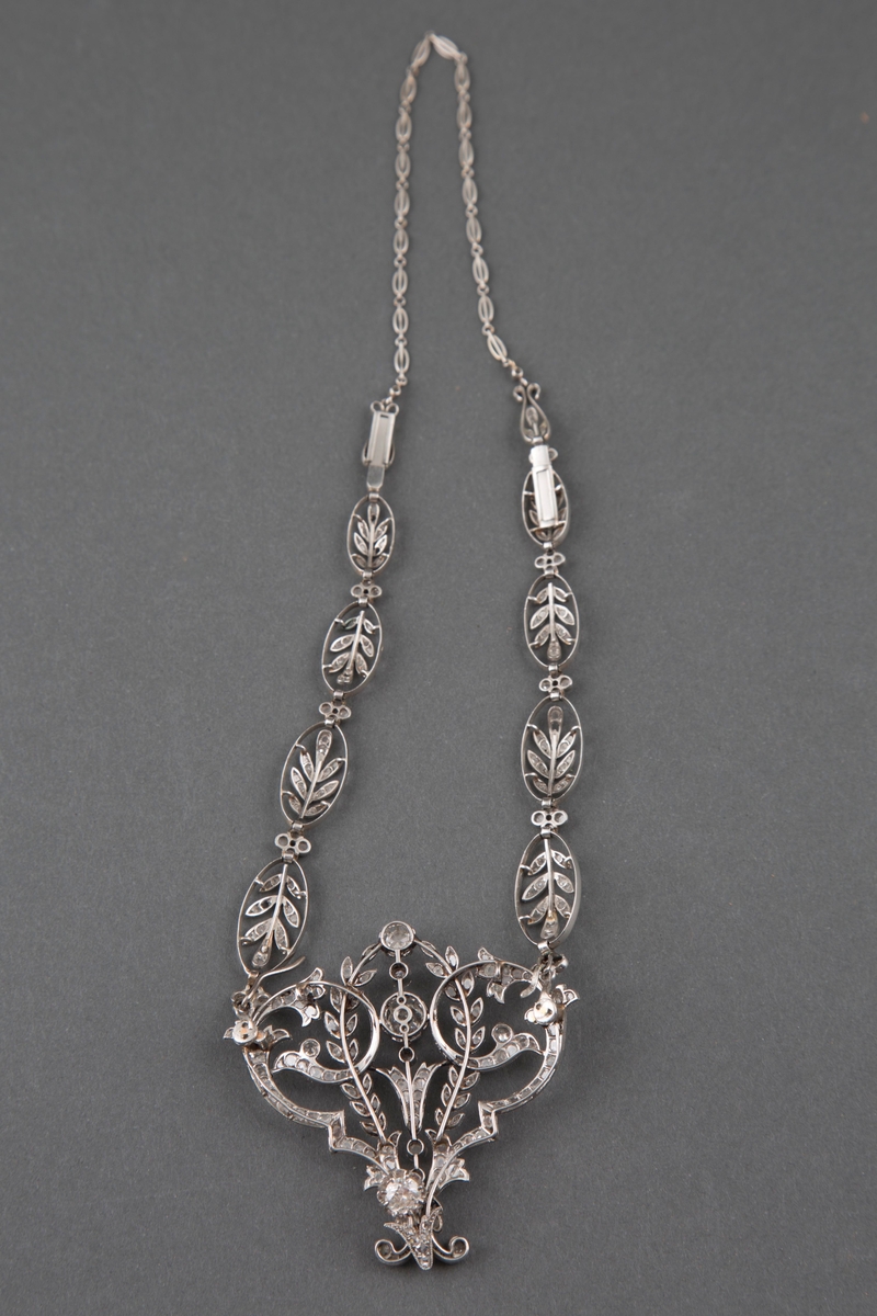 Halssmykke i platina og diamanter i originaleske. Kjedet består av ovaler med bladranker fylt med diamanter og små diamantblomster imellom. Anhenget er i hjerteform med blomster- og bladornamenter fylt med diamanter, samt runde innfattede diamanter i forskjellig størrelse.