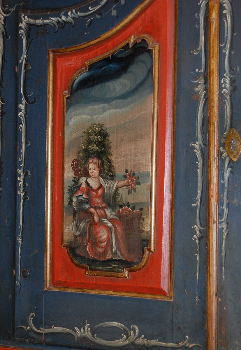 Skatoll av tre. Skapet har blå grunnfarge, og hvit og mørkeblå barokkmaling. På nederste del er det tre skuffer, med parvise håndtak laget av kobberlegering med gjennombrutt dekor. På øverste del av skapet er det to skapdører med fyllinger. I fyllingene er det malt dekor med motiv av to mennesker med blomster og fugl i hendene. Skapet har konveks koll, med to lister med gullbelagte kanter som ender i to volutter på midten. Mellom voluttene er det en gullbelagt kule med dekorative tverrsnitt. På den nedfellbare platen er det to fyllinger med profilerte lister, og malt dekor av landskapsmotiv.