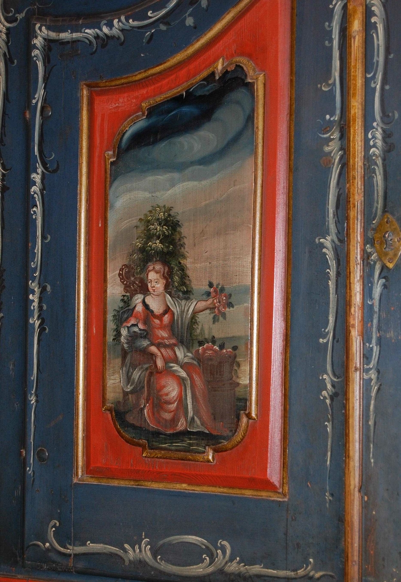 Skatoll av tre. Skapet har blå grunnfarge, og hvit og mørkeblå barokkmaling. På nederste del er det tre skuffer, med parvise håndtak laget av kobberlegering med gjennombrutt dekor. På øverste del av skapet er det to skapdører med fyllinger. I fyllingene er det malt dekor med motiv av to mennesker med blomster og fugl i hendene. Skapet har konveks koll, med to lister med gullbelagte kanter som ender i to volutter på midten. Mellom voluttene er det en gullbelagt kule med dekorative tverrsnitt. På den nedfellbare platen er det to fyllinger med profilerte lister, og malt dekor av landskapsmotiv.