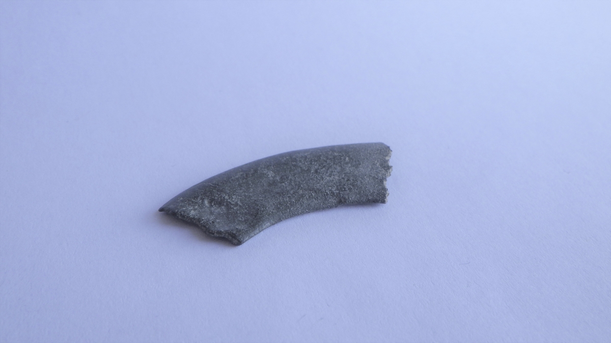 En avlang og buet metallplate med et tverrsnitt som buer svakt i kantene. Overflaten er glatt og baksiden mer ru. Gjenstandern har bruddflater i begge ender og ingen tegn til dekor. Dette er del av et rundt beslag, men med ukjent bruksområde.