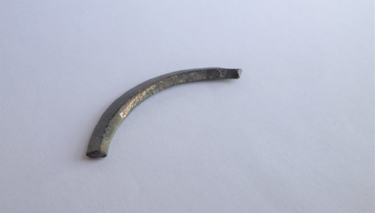 Et lengre og buet metallstykke med bruddflater i begge ender og trapesformet tverrsnitt. Gjenstanden er uten dekor, men støpt og filt. På undersiden er det forsatt noe støpespor som antyder at gjenstanden ikke er ferdig men et emne. Dette kan være en del av et emne til en ringspenne.