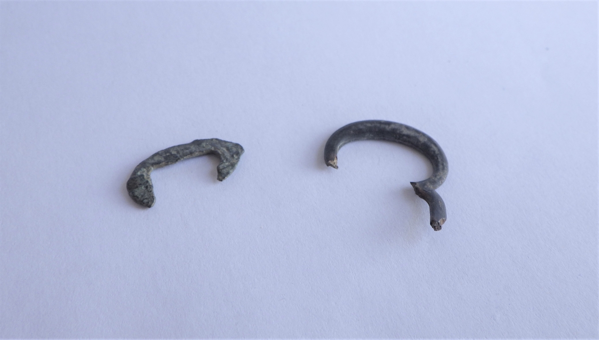 To små spenner av ulik størrelse og utforming.  Den minste er oval, med tynne, flate kanter og en rektangulær åpning i midten. Den større spennen har også oval utforming, oval indre rom og ser ut til å være en dobbeltspenne. Denne gjenstanden har også ovalt tverrsnitt på rammen. Ingen av de to spennene har bevart noe torn.