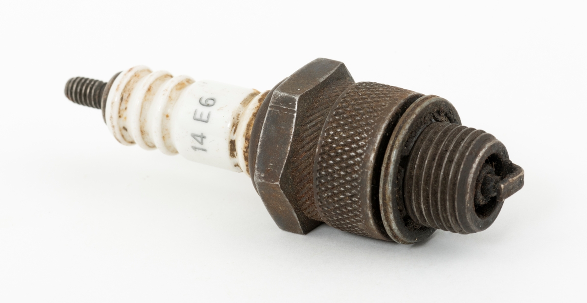 Tennplugg av merket «Prestolite». Dette er et nordamerikansk selskap som ble etablert i 1911 for å produsere bilelektriske artikler. Denne pluggen er drøyt 6,8 centimeter lang. Gjengetappen som ble innskrudd øverst i motorens sylinder har en diameter på 1,4 centimeter. Pluggen veier 54 gram. Det kvite porselenselementet som isolerer den øvre delen av pluggen er merket med fabrikantnavnet «prestolite» og tall- og bokstavkoden «14 E6». Denne koden angir sannsynligvis varmeverdien til pluggen. Tennpluggens oppgave er å omdanne høyspenning fra bensinmotorens tennspole til en gnist i forbrenningskammeret øverst i sylinderen. Tennpluggen ble avlevert til museet i ei kasse som inneholdt materiell fra ei smie.