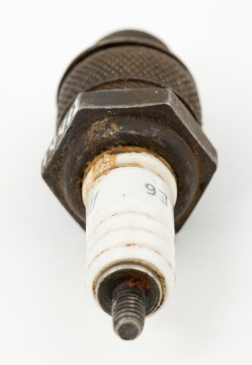 Tennplugg av merket «Prestolite». Dette er et nordamerikansk selskap som ble etablert i 1911 for å produsere bilelektriske artikler. Denne pluggen er drøyt 6,8 centimeter lang. Gjengetappen som ble innskrudd øverst i motorens sylinder har en diameter på 1,4 centimeter. Pluggen veier 54 gram. Det kvite porselenselementet som isolerer den øvre delen av pluggen er merket med fabrikantnavnet «prestolite» og tall- og bokstavkoden «14 E6». Denne koden angir sannsynligvis varmeverdien til pluggen. Tennpluggens oppgave er å omdanne høyspenning fra bensinmotorens tennspole til en gnist i forbrenningskammeret øverst i sylinderen. Tennpluggen ble avlevert til museet i ei kasse som inneholdt materiell fra ei smie.