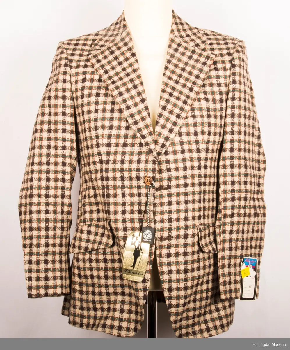 Kavaler dressjakke mønstrete ullstoff. Sennepsgult syntetisk silkefor. Jakka har skulderputer, store klaffer på lommene, 2 splitter bak.   Den er knept med 2 plastknapper midt framme.  1 pynteknapp på hvert erme.  Store slag.