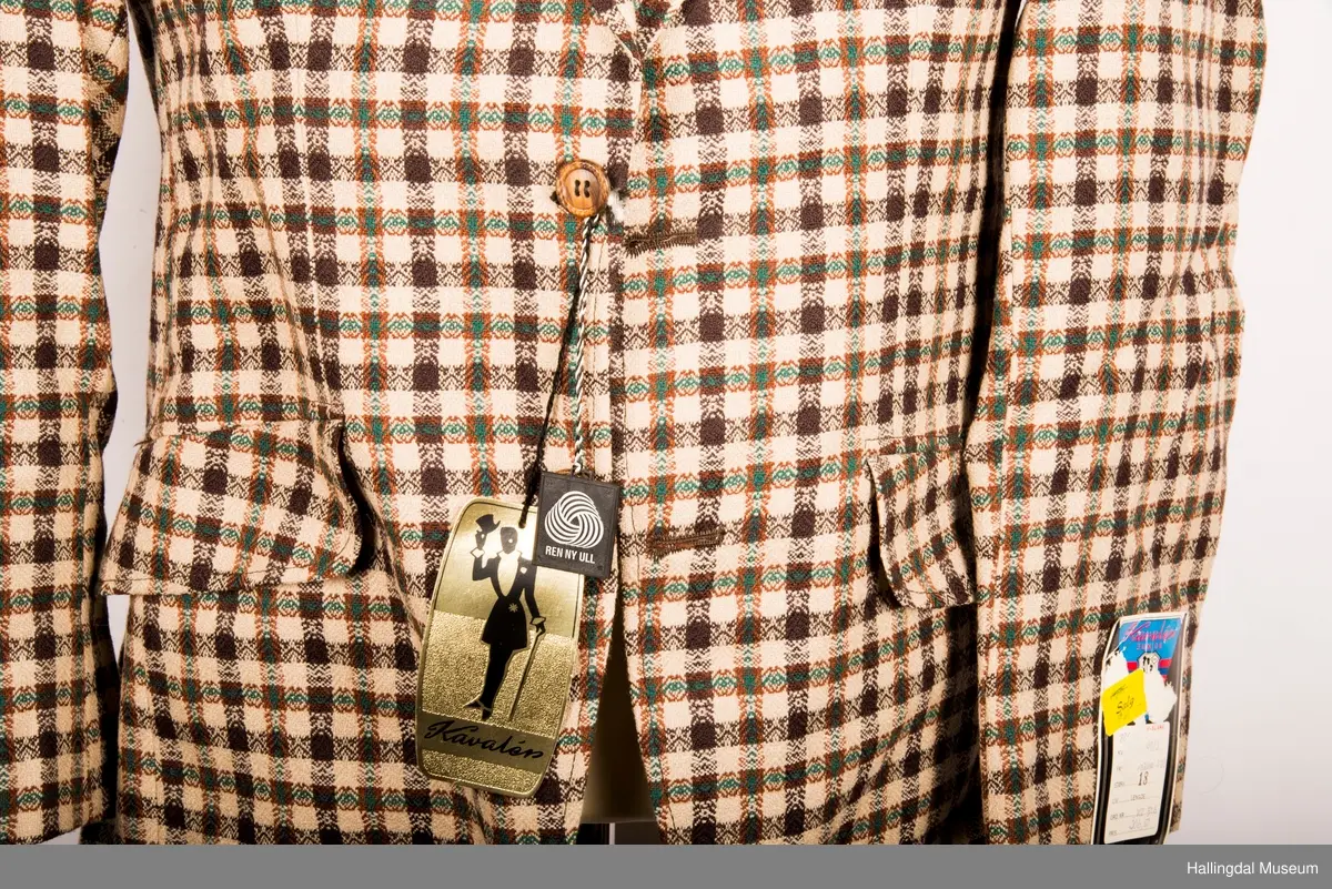 Kavaler dressjakke mønstrete ullstoff. Sennepsgult syntetisk silkefor. Jakka har skulderputer, store klaffer på lommene, 2 splitter bak.   Den er knept med 2 plastknapper midt framme.  1 pynteknapp på hvert erme.  Store slag.