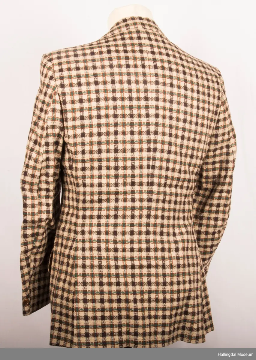 Kavaler dressjakke mønstrete ullstoff. Sennepsgult syntetisk silkefor. Jakka har skulderputer, store klaffer på lommene, 2 splitter bak.   Den er knept med 2 plastknapper midt framme.  1 pynteknapp på hvert erme.  Store slag.