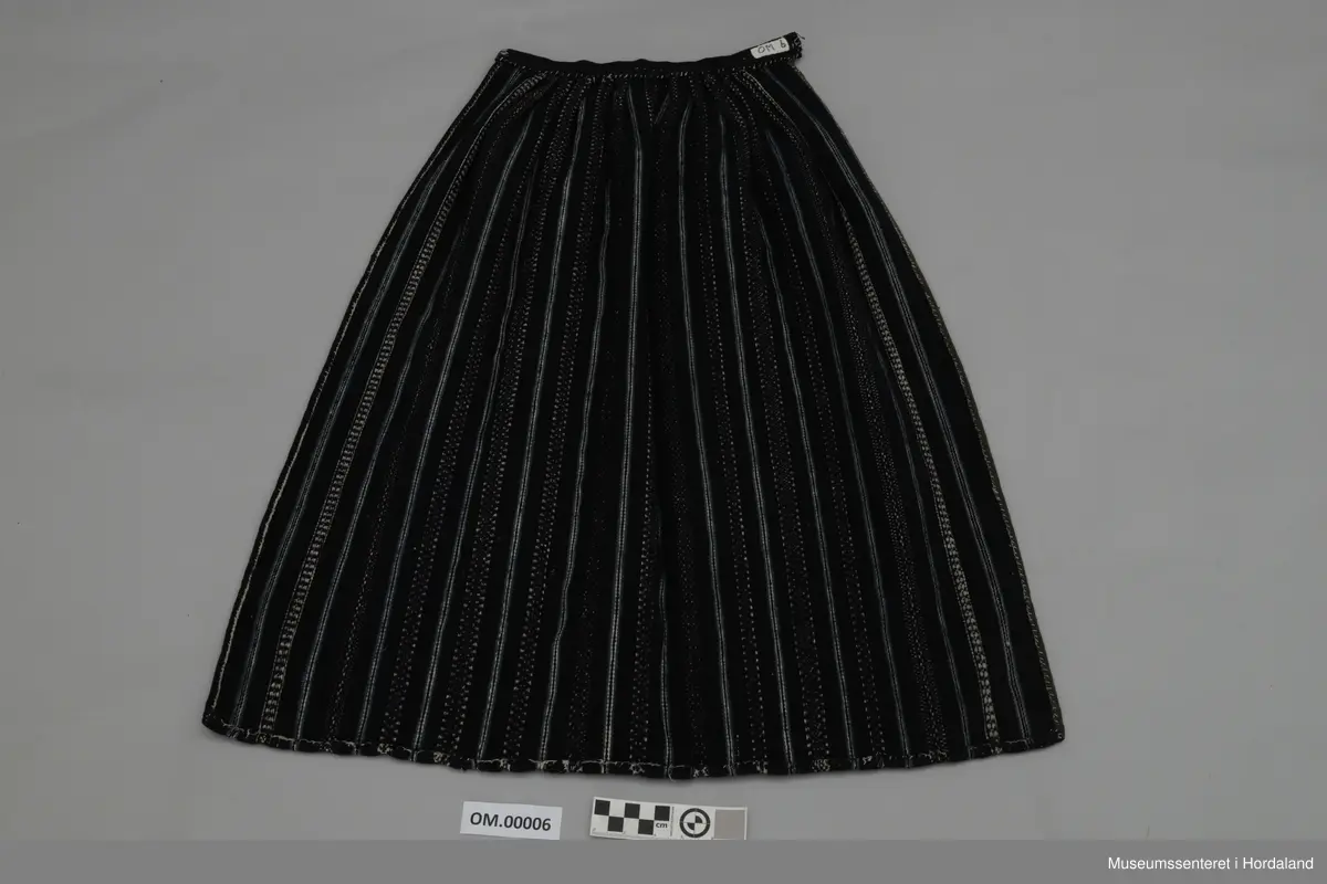 Mørkeblått, vove, stripete forkle i ull. Stripene er kvite og lyseblå. Linning med ein ring i kvar ende for feste av forkleband. Smalt, glatt stykke framme med seks folder på kvar side.