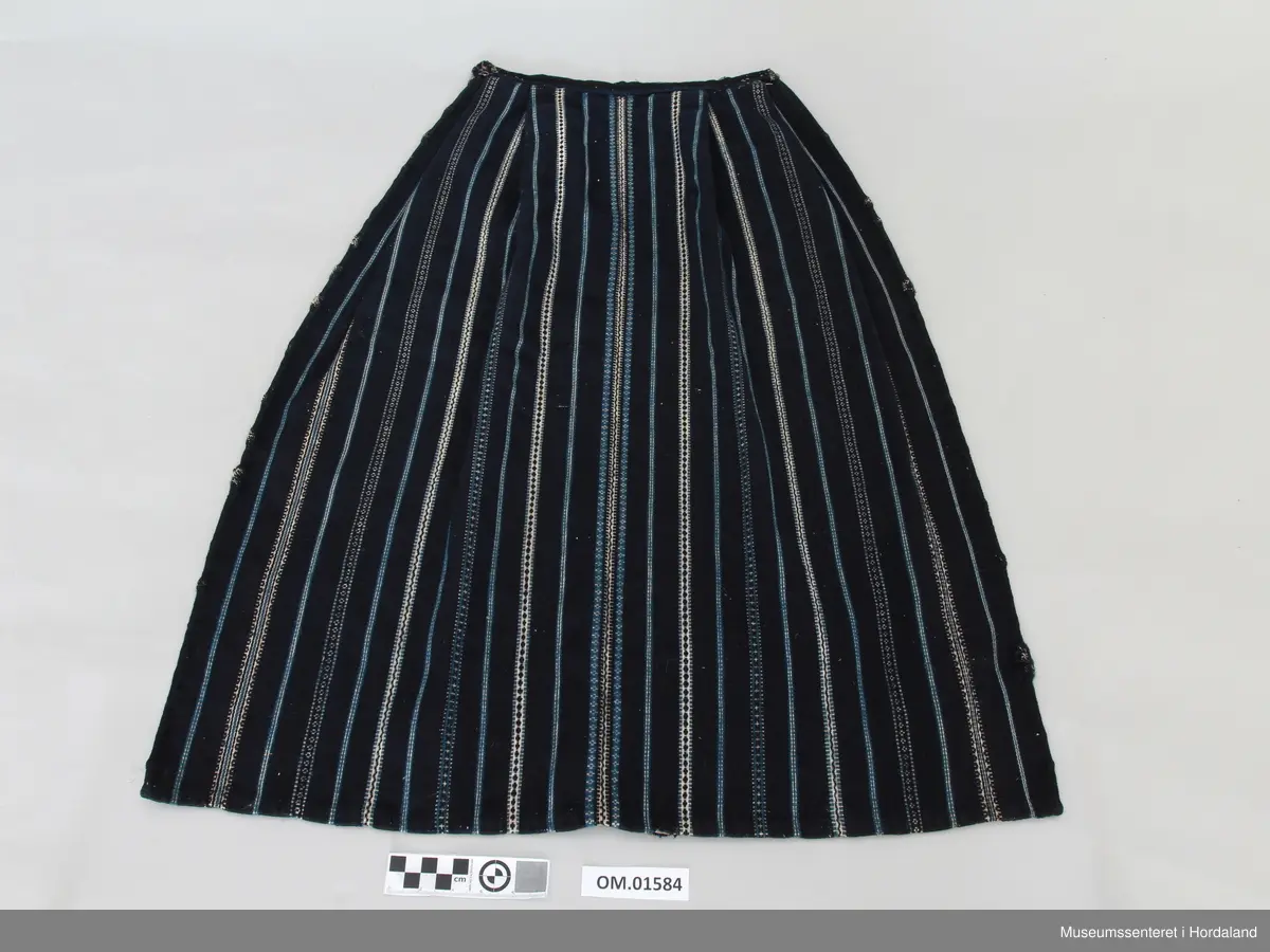 Mørkeblått, vove ullforkle med striper i kvitt og mellomblått. Linning. Glatt stykke midt framme, to folder på kvar side. Ei hekte til attlat.
