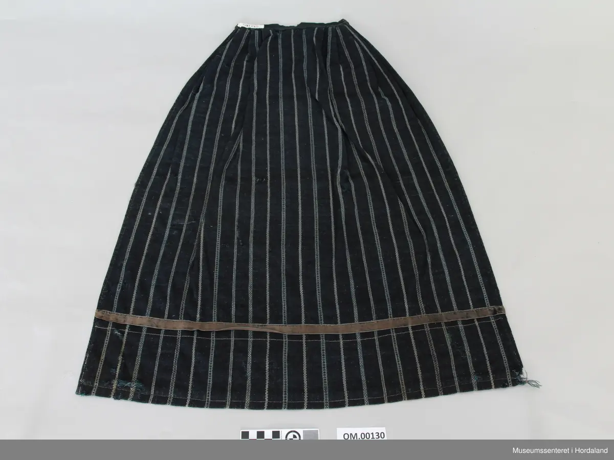 Mørkeblått, stripete vove forkle i ull. Striper i kvitt og blått mønster. Linning. Glatt stykke midt framme, tre folder på kvar side. Brunt fløyelsband nede. Skoning på baksiden med mønstra bomullsstoff og saum som syner på framsida. Forkle er smalt oppe. Holete.