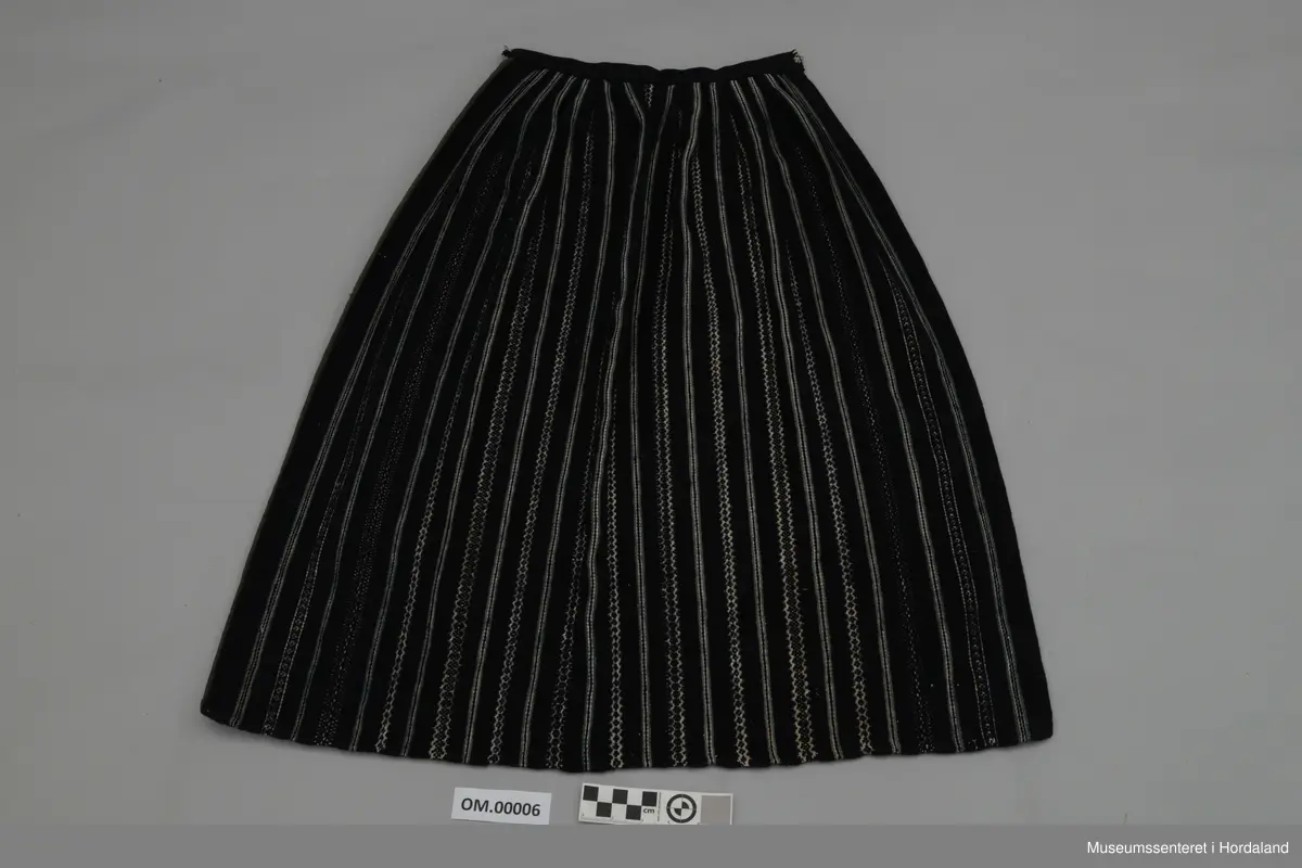 Mørkeblått, vove, stripete forkle i ull. Stripene er kvite og lyseblå. Linning med ein ring i kvar ende for feste av forkleband. Smalt, glatt stykke framme med seks folder på kvar side.