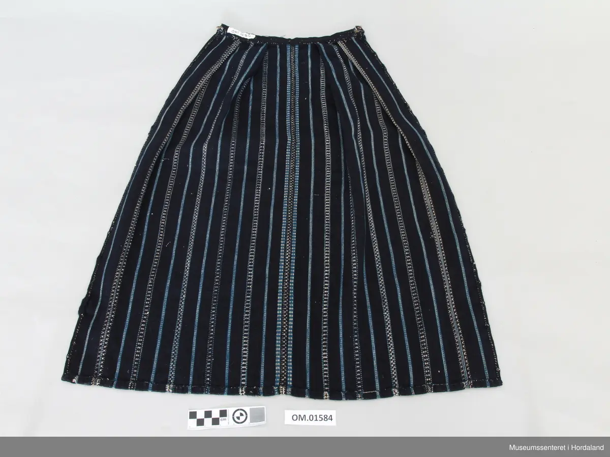 Mørkeblått, vove ullforkle med striper i kvitt og mellomblått. Linning. Glatt stykke midt framme, to folder på kvar side. Ei hekte til attlat.