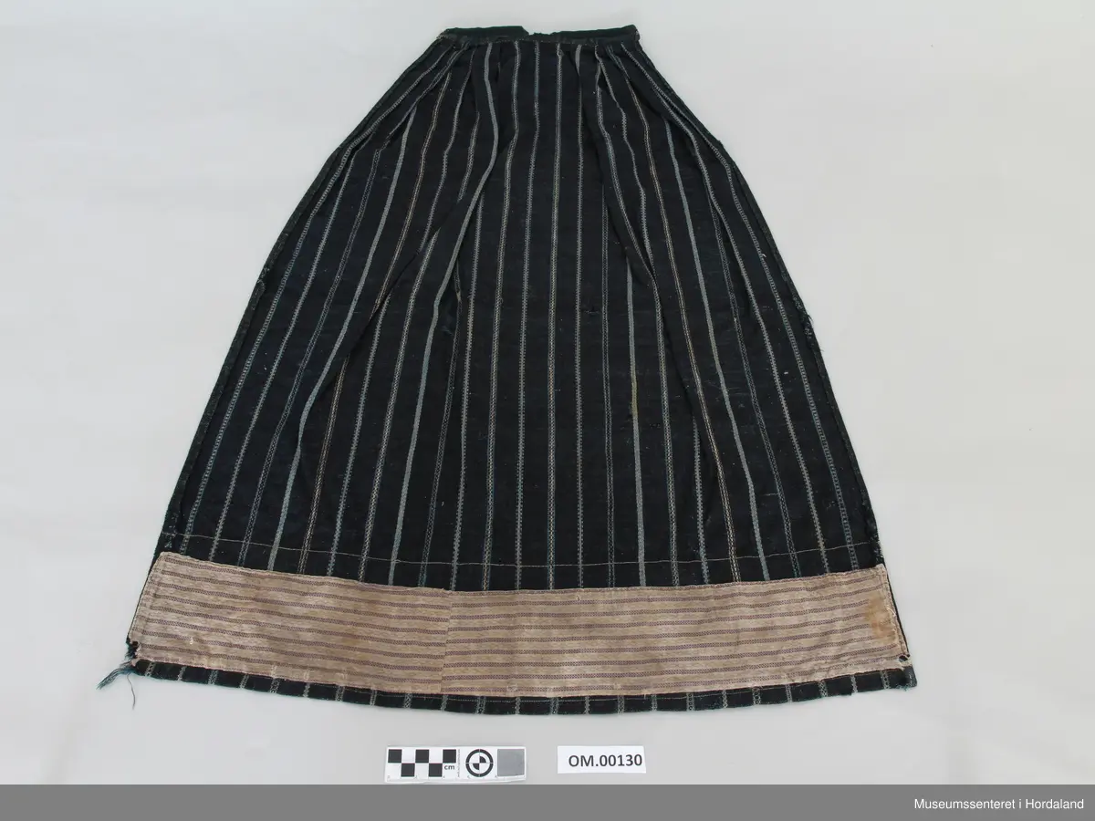 Mørkeblått, stripete vove forkle i ull. Striper i kvitt og blått mønster. Linning. Glatt stykke midt framme, tre folder på kvar side. Brunt fløyelsband nede. Skoning på baksiden med mønstra bomullsstoff og saum som syner på framsida. Forkle er smalt oppe. Holete.