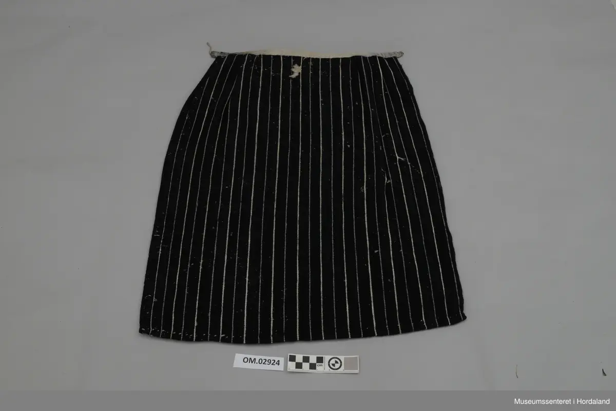 Mørkeblått forkle med kvite og lyselilla, smale og jamne striper. Linning av silkeband. To ringar med hyssing. Framme er det glatt parti og to folder på kvar side.