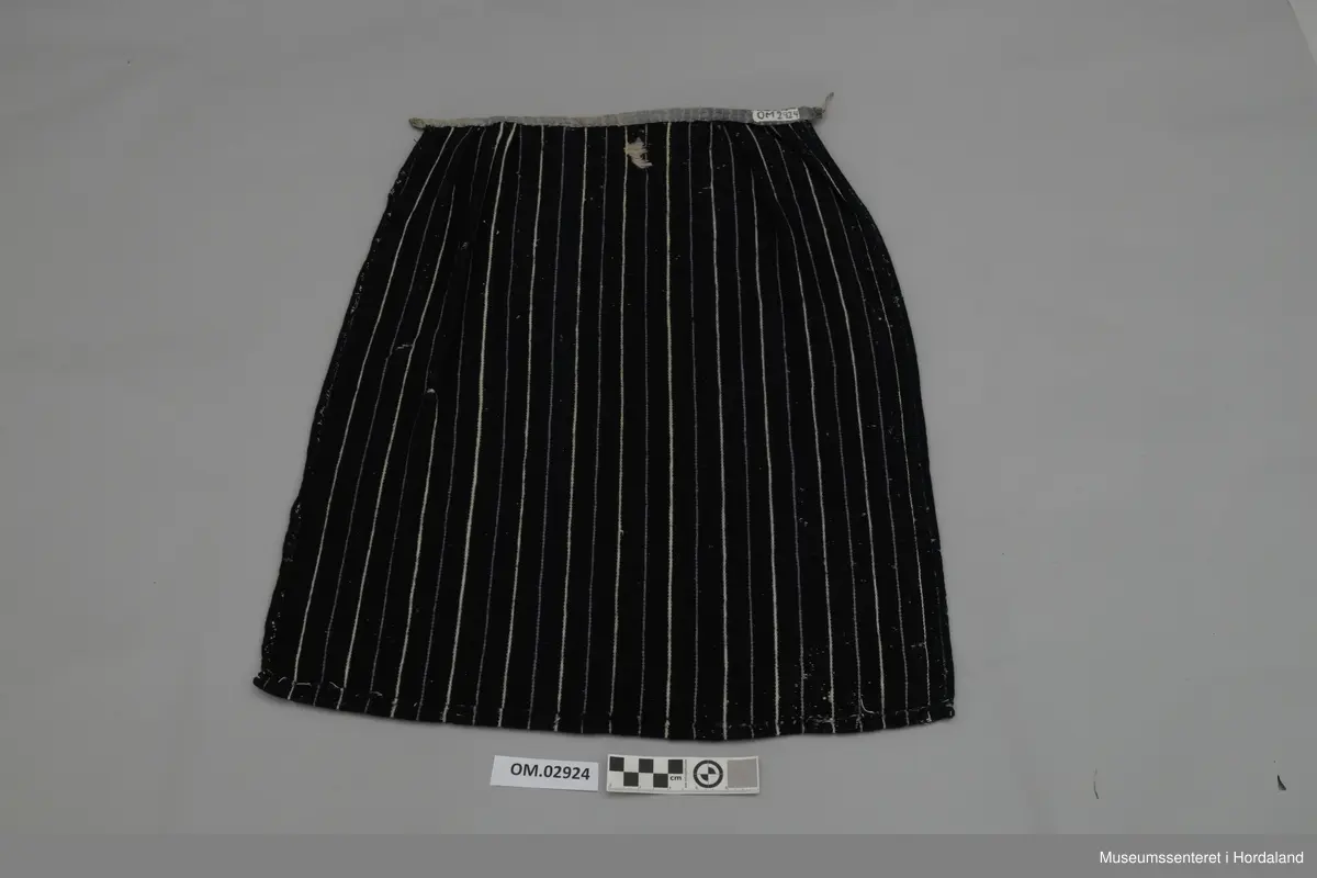 Mørkeblått forkle med kvite og lyselilla, smale og jamne striper. Linning av silkeband. To ringar med hyssing. Framme er det glatt parti og to folder på kvar side.