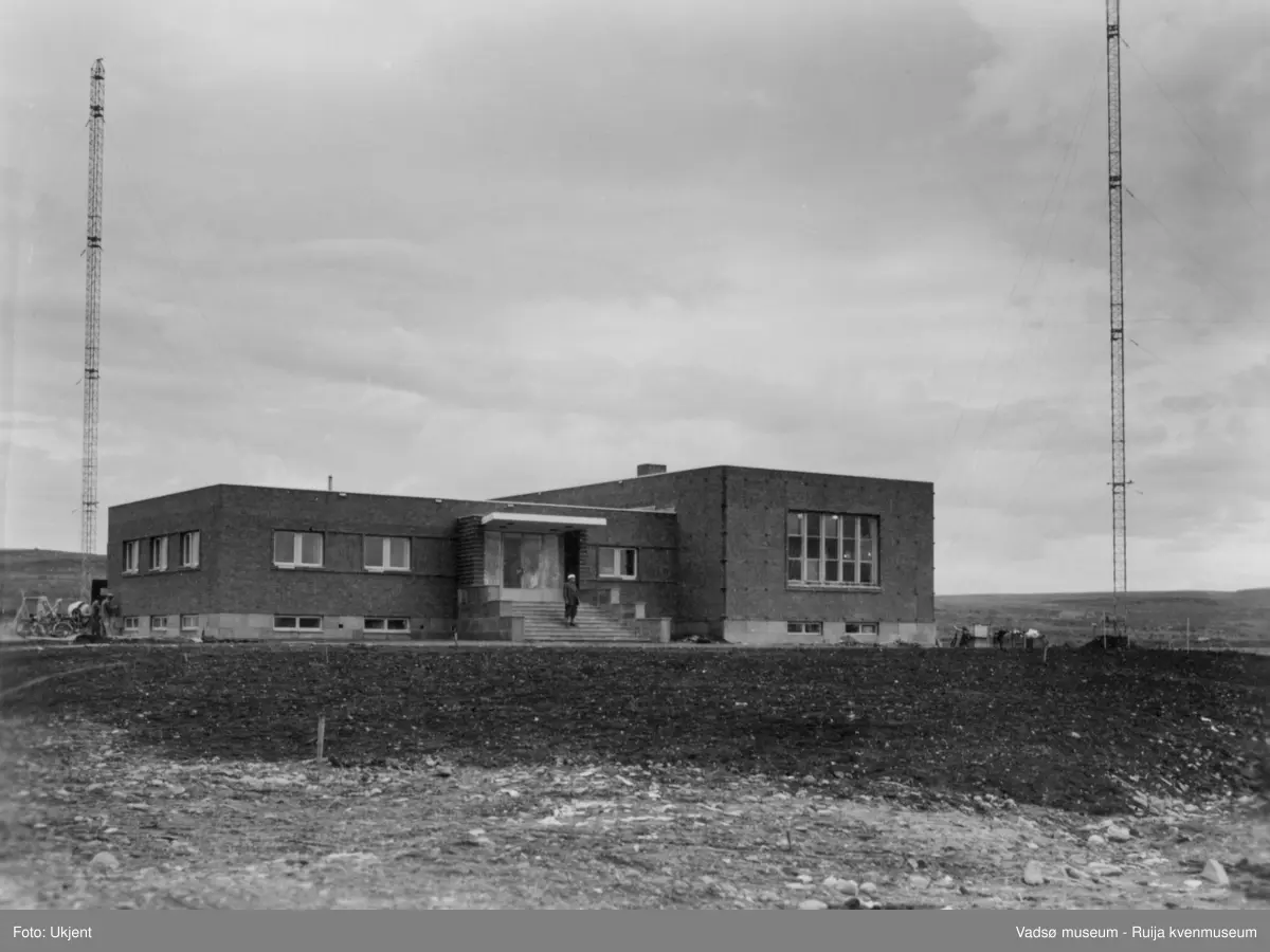 NRK-bygget i Vadsø. Bildet er tatt mot nordøst. Vi ser NRK-bygget med to radiomaster. På trappa står en mann. På vestsiden av bygningen ser vi to mann i arbeid på vestveggen. Ved siden av står en sementblandemaskin og et par sykler. Området i front - i syd av bygningen - er nyplanert. På østsiden av bygget står en sykkel støttet opp mot et par kasser. 
Finnmark kringkaster (NRK-bygget) i Vadsø, byggeår 1949. Arkitekt Henrik L. Kjær. Bygget har bærende betongvegger og fasadene er lagt med teglstein og grunnmuren er belagt med skiferplater. (Kilde: Arkitekturguide for Nord-Norge og Svalbard)