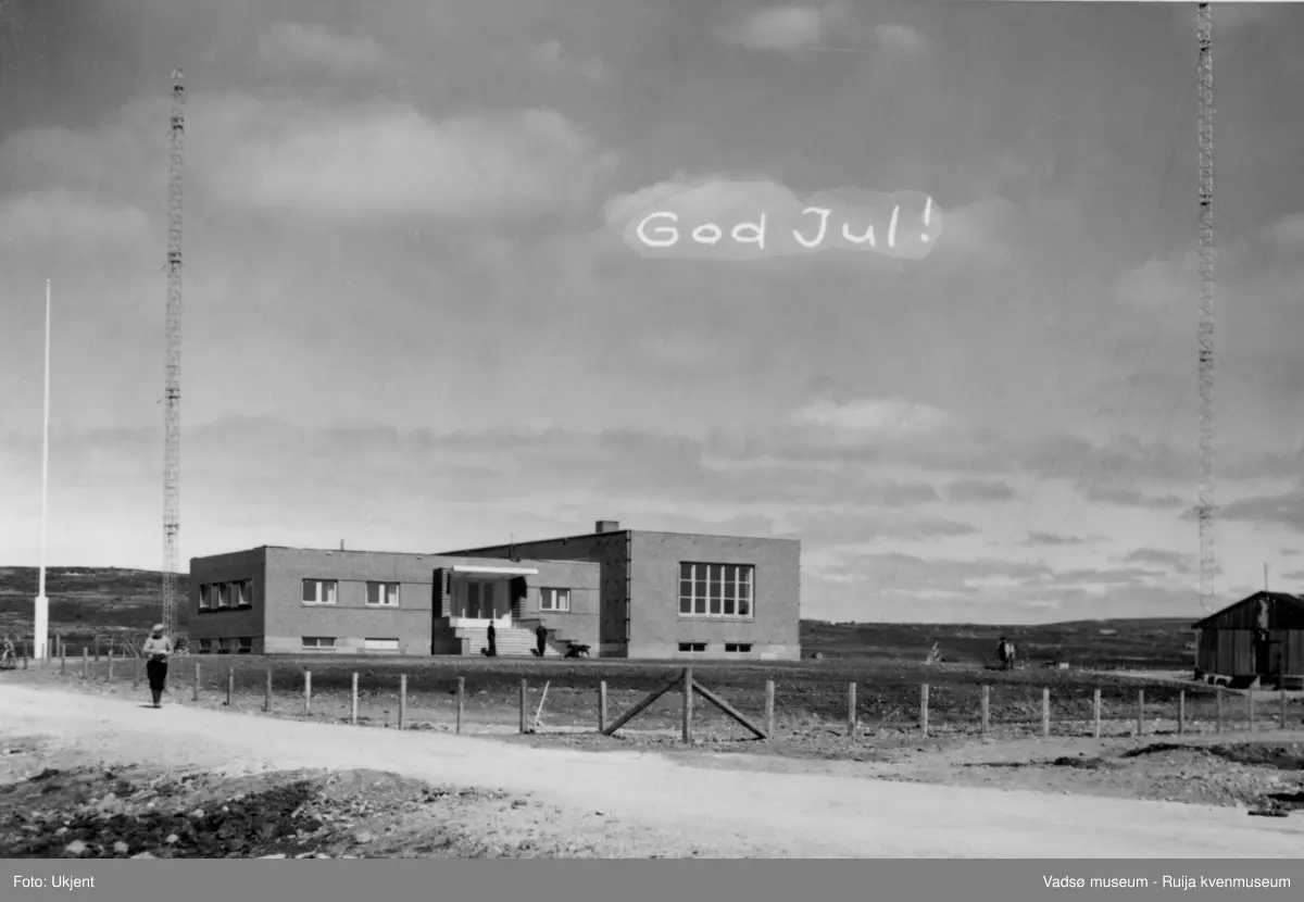 NRK-bygget i Vadsø. Bildet er tatt mot nordøst. Vi ser NRK-bygget med to radiomaster og flaggstang. Nedenfor trappa står det to menn. Det er satt opp et gjerde rundt bygningen og området. I forkant av bildet, på vestsiden av bygningen, ser vi en mann komme spaserende ned langs Fosseveien. På østsiden av bygget står en brakke.  Midt på bildet er det skrevet en hilsen for hånd; God Jul!
Finnmark kringkaster (NRK-bygget) i Vadsø, byggeår 1949. Arkitekt Henrik L. Kjær. Bygget har bærende betongvegger og fasadene er lagt med teglstein og grunnmuren er belagt med skiferplater. (Kilde: Arkitekturguide for Nord-Norge og Svalbard)