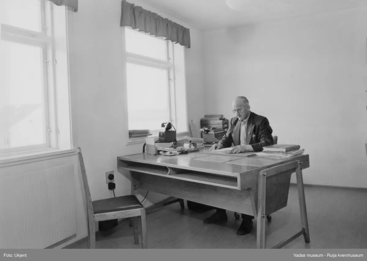 NRK-bygget i Vadsø. Et kontor med to vinduer, en radiator under det ene vinduet. En mann med briller sitter ved et skrivebord.  På skrivebordet ser vi en telefon på et stativ samt en del kontorutstyr. På et lite bord i hjørnet av rommet står en skrivemaskin og noen arkivbokser. 
Finnmark kringkaster (NRK-bygget) i Vadsø, byggeår 1949. Arkitekt Henrik L. Kjær. Bygget har bærende betongvegger og fasadene er lagt med teglstein og grunnmuren er belagt med skiferplater. (Kilde: Arkitekturguide for Nord-Norge og Svalbard)