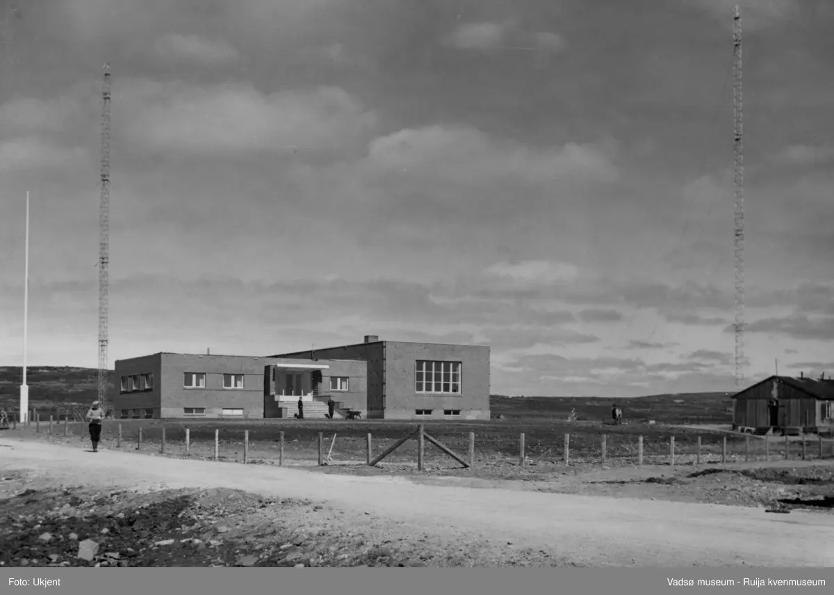 NRK-bygget i Vadsø. Bildet er tatt mot nordøst. Vi ser NRK-bygget med to radiomaster og flaggstang. Nedenfor trappa står det to menn. Det er satt opp et gjerde rundt bygningen og området. I forkant av bildet, på vestsiden av bygningen, ser vi en mann komme spaserende ned langs Fosseveien. På østsiden av bygget står en brakke.  
Finnmark kringkaster (NRK-bygget) i Vadsø, byggeår 1949. Arkitekt Henrik L. Kjær. Bygget har bærende betongvegger og fasadene er lagt med teglstein og grunnmuren er belagt med skiferplater. (Kilde: Arkitekturguide for Nord-Norge og Svalbard)