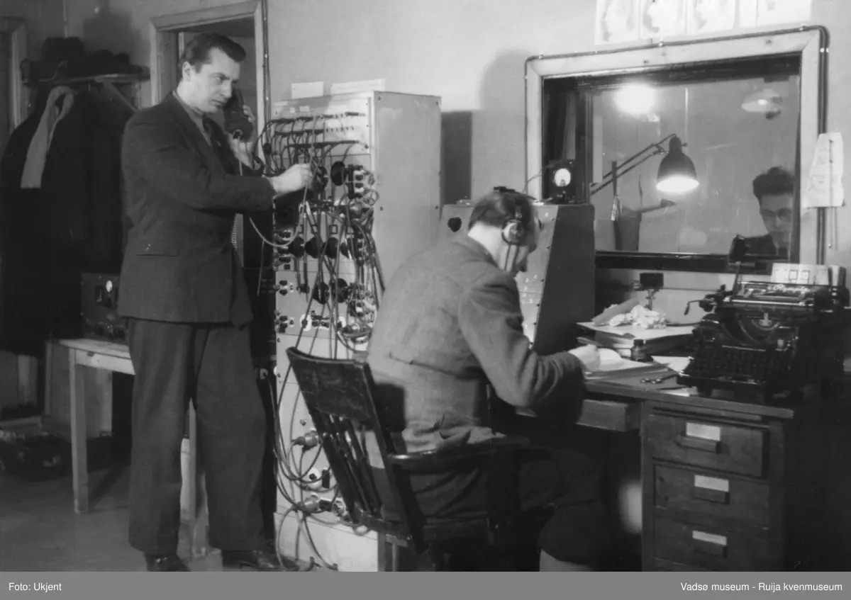 Finnmark Kringkaster i Vadsø, mars 1949. I det nærmeste rommet, kontrollrommet,  står en mann ved et apparat og ved skrivebord sitter en mann med øretelefoner. På skrivebordet ser vi bl.a. en skrivemaskin. Bak en glassrute, i et tilstøtende rom, studio, sitter en mann med briller. 
Bildet er også gjengitt i boka "Fra djevelmaster til Hallo fiskeflåten" av Bård Gudmund Hansen, med tekst: "Erling Grønvigh og Nils Fr. Jørgensen prøver å få Ivar Balsvik (bak ruta) på lufta, mars 1949."