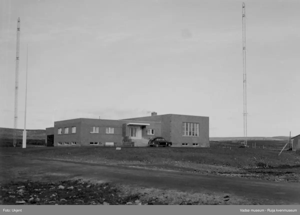 NRK-bygget i Vadsø. Bildet er tatt mot nordøst. Vi ser NRK-bygget med to radiomaster. På vestsiden av bygget står en hvit flaggsang. Nedenfor trappa står en sort personbil parkert. I høyre billedkant ser vi deler av en brakke. 
Finnmark kringkaster (NRK-bygget) i Vadsø, byggeår 1949. Arkitekt Henrik L. Kjær. Bygget har bærende betongvegger og fasadene er lagt med teglstein og grunnmuren er belagt med skiferplater. (Kilde: Arkitekturguide for Nord-Norge og Svalbard)