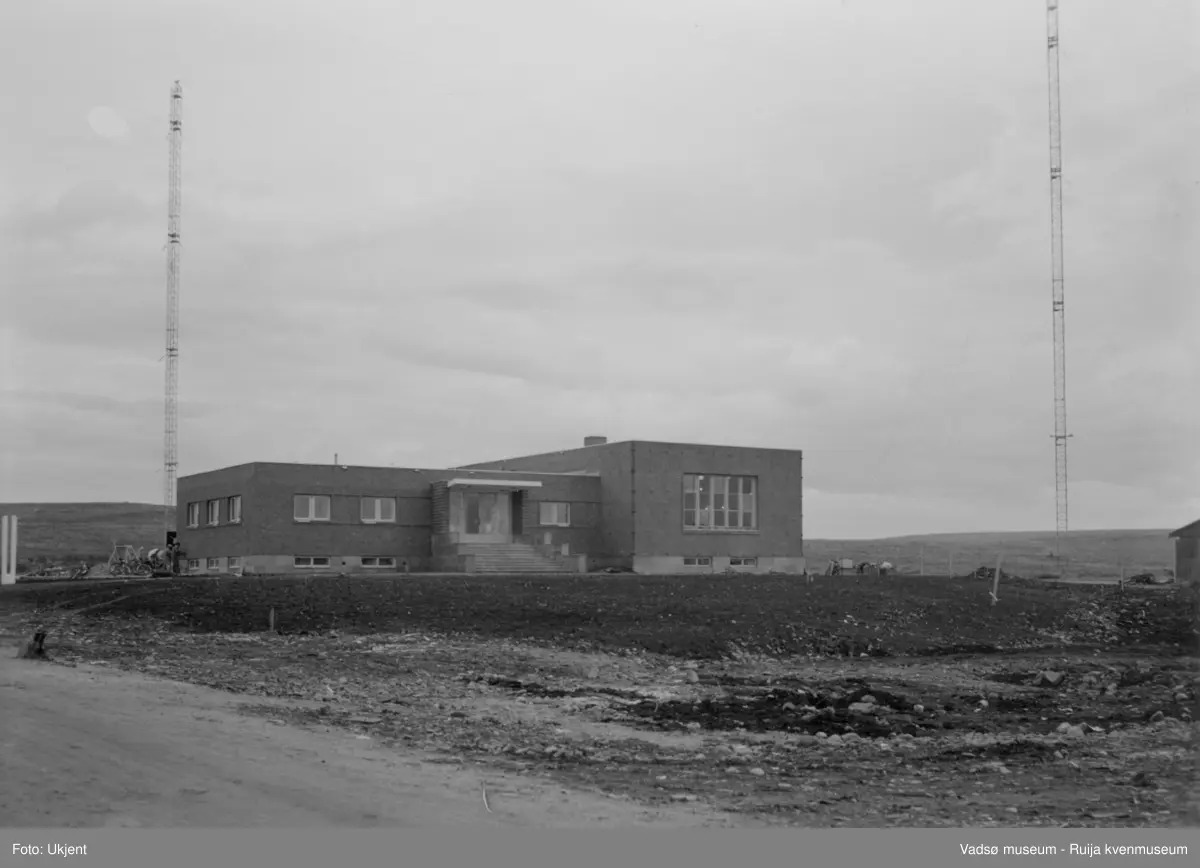 NRK-bygget i Vadsø. Bildet er tatt mot nordøst. Vi ser NRK-bygget med to radiomaster. På vestsiden av bygningen ser vi deler av flaggstanga og to mann i arbeid på vestveggen. Ved siden av står en sementblandemaskin og et par sykler. Området i front - i syd av bygningen - er nyplanert. På østsiden av bygget står en sykkel støttet opp mot noen kasser. Helt i høyre billedkant ser vi hjørnet av en brakke. 
Finnmark kringkaster (NRK-bygget) i Vadsø, byggeår 1949. Arkitekt Henrik L. Kjær. Bygget har bærende betongvegger og fasadene er lagt med teglstein og grunnmuren er belagt med skiferplater. (Kilde: Arkitekturguide for Nord-Norge og Svalbard)