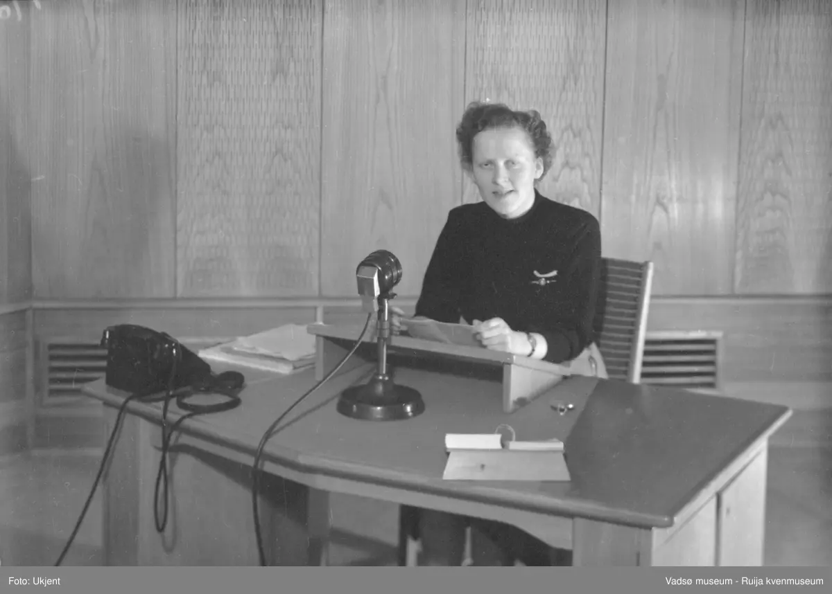 NRK-bygget i Vadsø. Ved skrivebordet i studio sitter programsekretær Anna Dahl Johnsen. På skrivebordet ser vi en telefon, en mikrofon, en kalender og noen dokumenter.  
Finnmark kringkaster (NRK-bygget) i Vadsø, byggeår 1949. Arkitekt Henrik L. Kjær. Bygget har bærende betongvegger og fasadene er lagt med teglstein og grunnmuren er belagt med skiferplater. (Kilde: Arkitekturguide for Nord-Norge og Svalbard og "Fra djevelmaster til Hallo fiskeflåten" av Bård Gudmund Hansen)
Kil