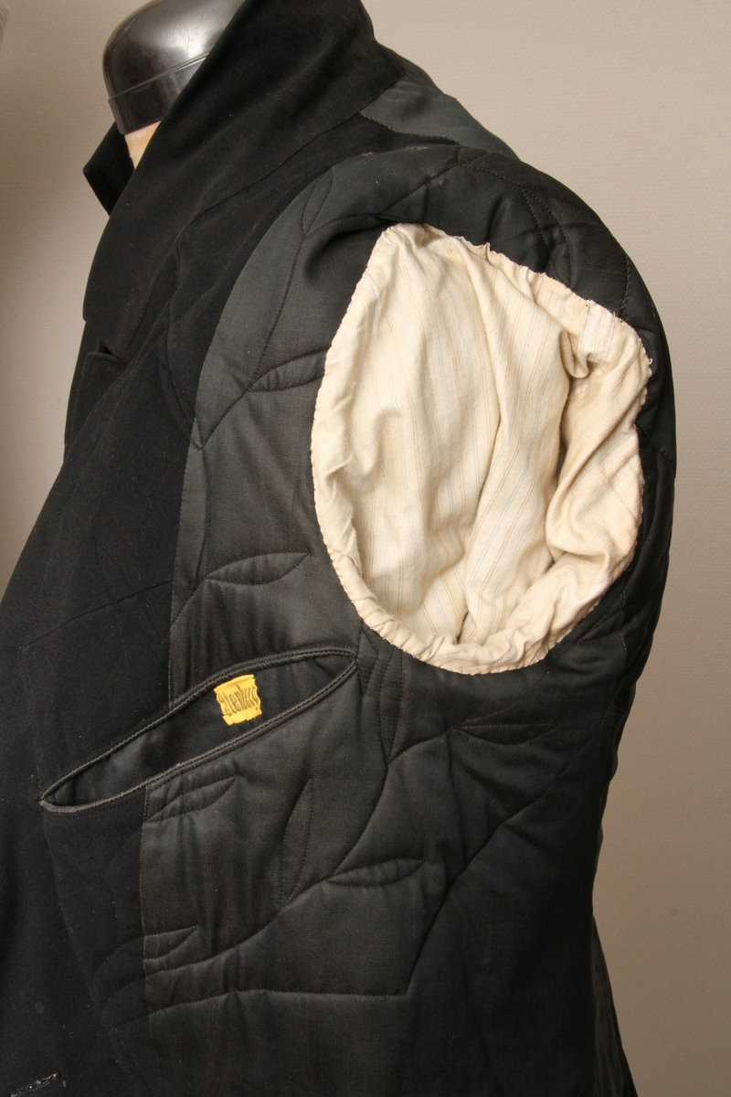 Lang jakke sydd i svart, blankt ullstoff. Krage med slag, dobbeltspent med trekte knapper. Søm rundt hele livlinja, påsydd to store flak, 50 cm lange. Lang splitt bak, en knapp påsydd bak på hvert flak. To innerlommer, samt to stikklommer i skjørteflakene. Bryststykkene er vattert og har maskinsømstikninger innvendig. Resten av jakka er sydd for hånd. 8 cm bred kant på ermene, pluss to mindre trekte knapper. Merket med trykt bånd "Stenberg". 