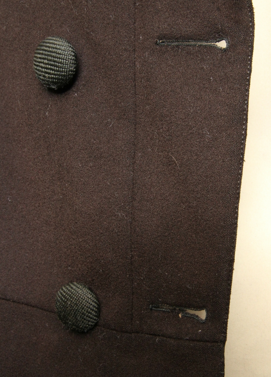Lang jakke sydd i brunt, blankt ullstoff. Krage med slag, dobbeltspent med trekte knapper. Søm rundt hele livlinja, med unntak av ryggstykkene som er hele. Påsydd to store flak, 55 cm lange. Lang splitt bak, en knapp påsydd bak på hvert flak. En innerlomme, samt to stikklommer i skjørteflakene. Hele jakka er sydd for hånd. Jakka er godt brukt, og det er sydd på lapper i nakken, nederst i ermeforet og i livlinja. 