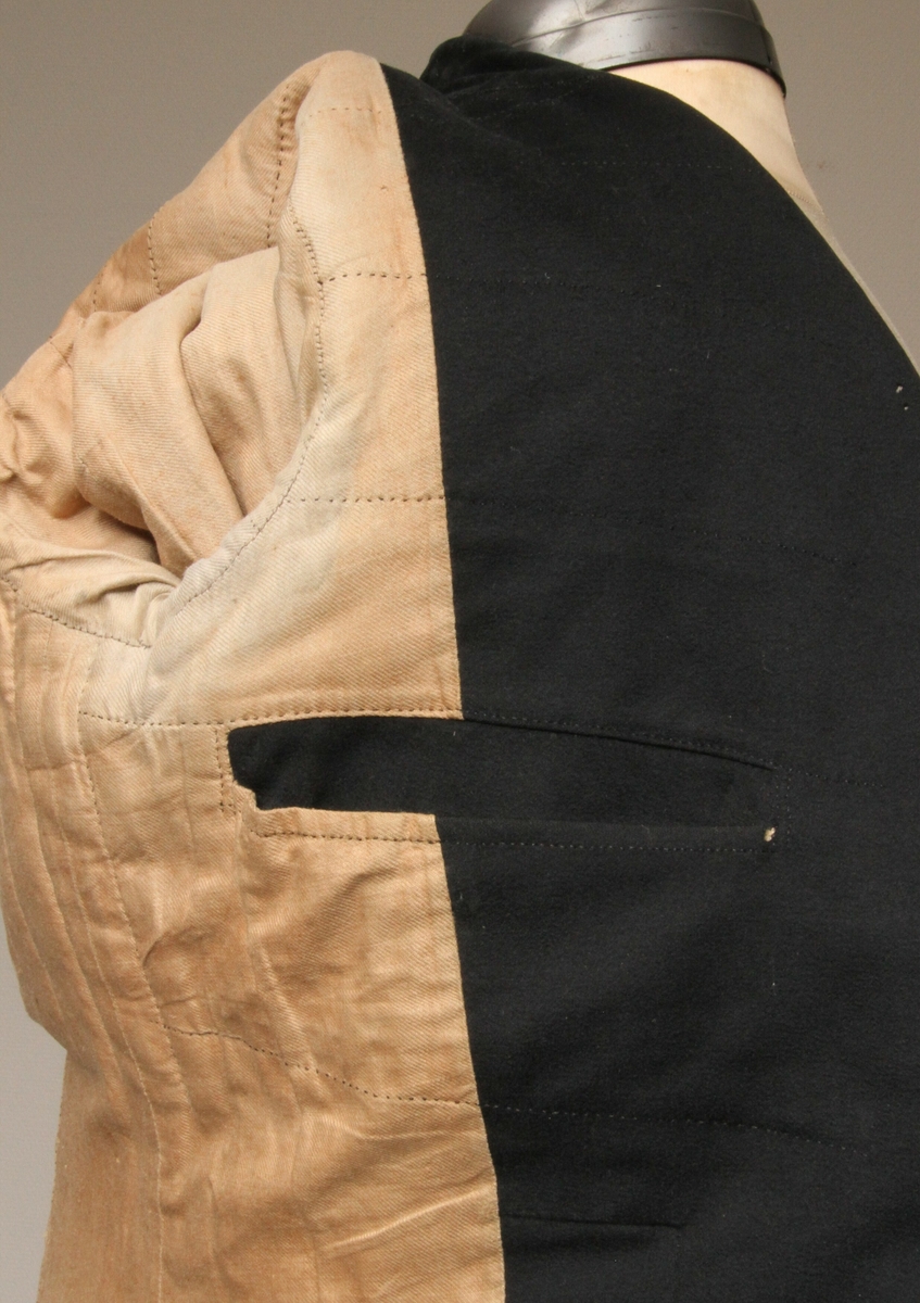 Jakke sydd i svart, blankt ullstoff. Krage med slag, dobbeltspent med trekte knapper. Påsydd to store flak bak, 38 cm lange midt bak. Lang splitt, med en knapp påsydd bak på hvert flak både øverst og nederst. To innerlommer, samt to stikklommer i skjørteflakene. Sidestykkene er vattert og hele jakka er foret. En rekke stikninger innvendig. Hele jakka er sydd for hånd med brun lintråd. 